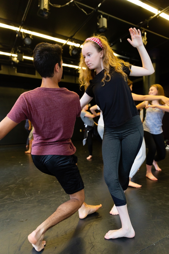 Two drama students face each other, linking arms in mid-movement during an acting studio class